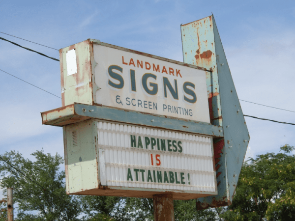 large antique sign, rusted, for the landmark sign company. it has a message board that reads "happiness is attainable." this is incongruous due to the state of the sign.